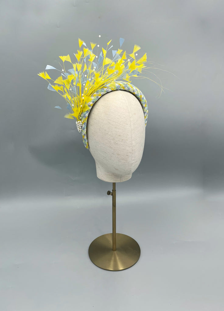 Bespoke millinery headband featuring a braided pastel blue and yellow base, adorned with a dramatic spray of yellow and pale blue feathers, finished with pearl embellishment.